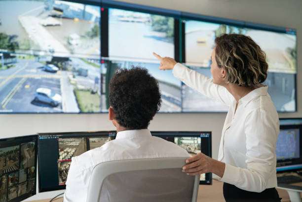 Rear view of female and male security coworkers discussing while looking and pointing at video wall in control room